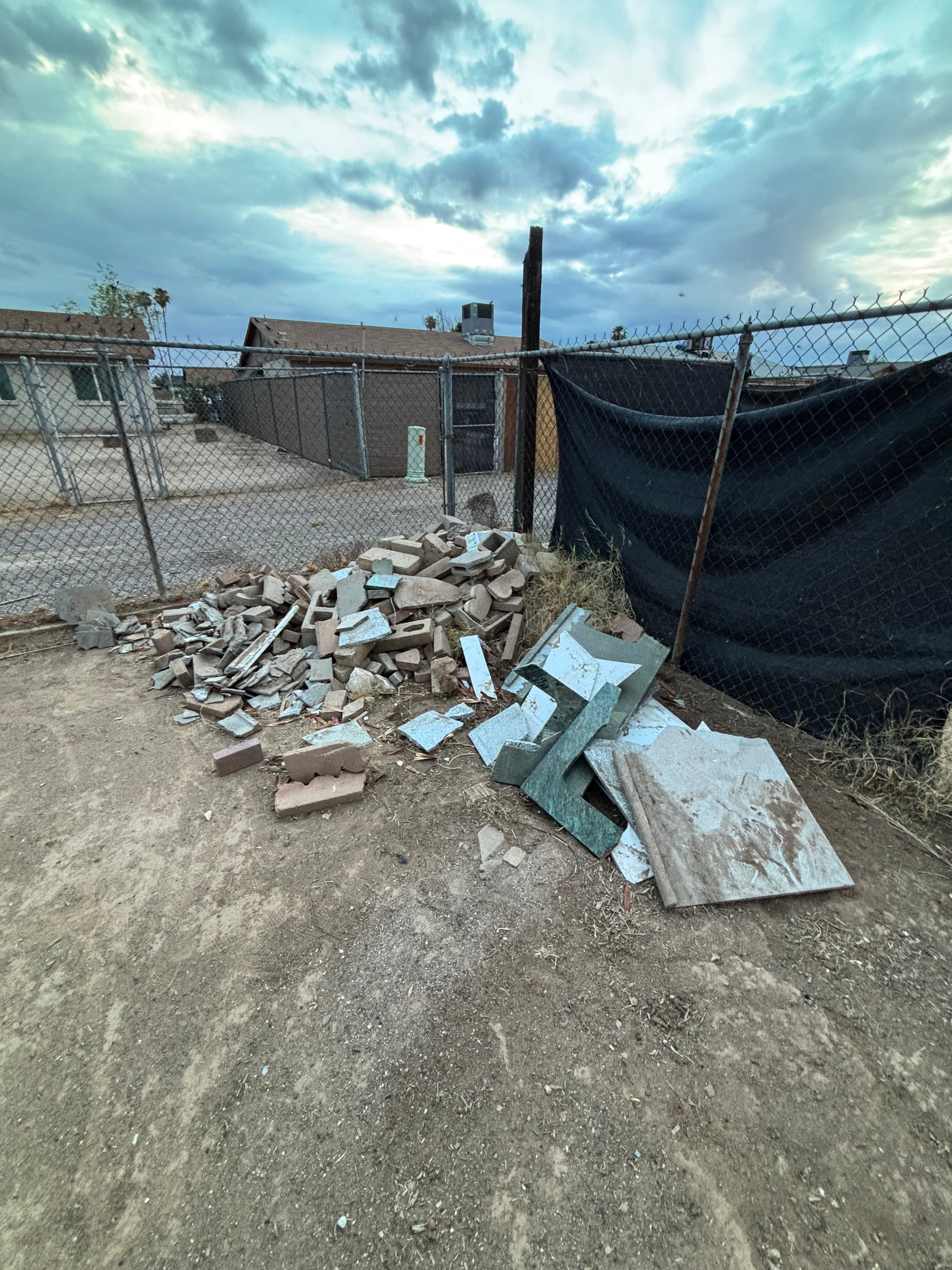 Broken bricks and tiles piled on dirt beside a chain-link fence under cloudy skies.