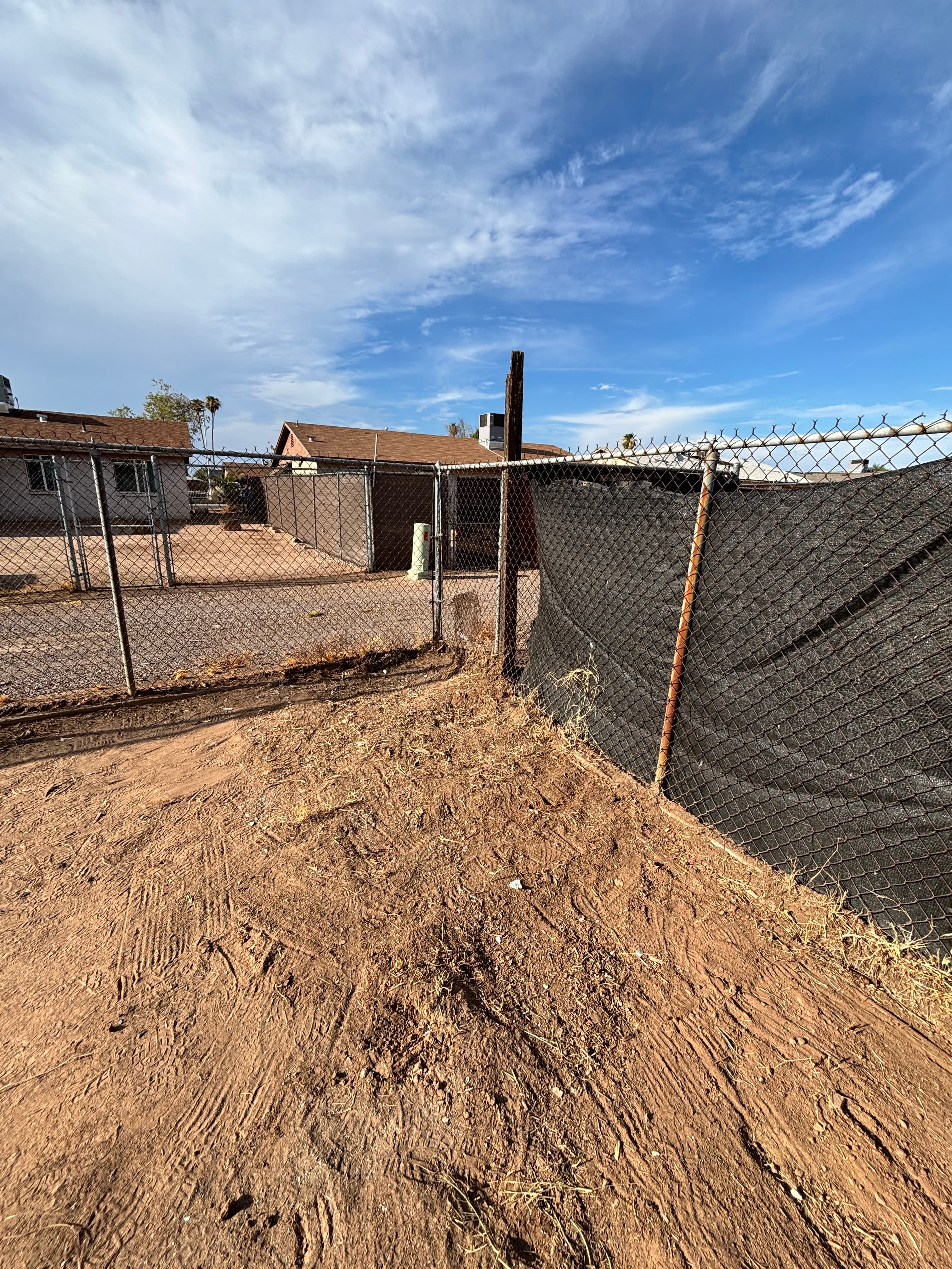 Tire tracks on dirt lead to a chain-link fence and houses under blue skies.