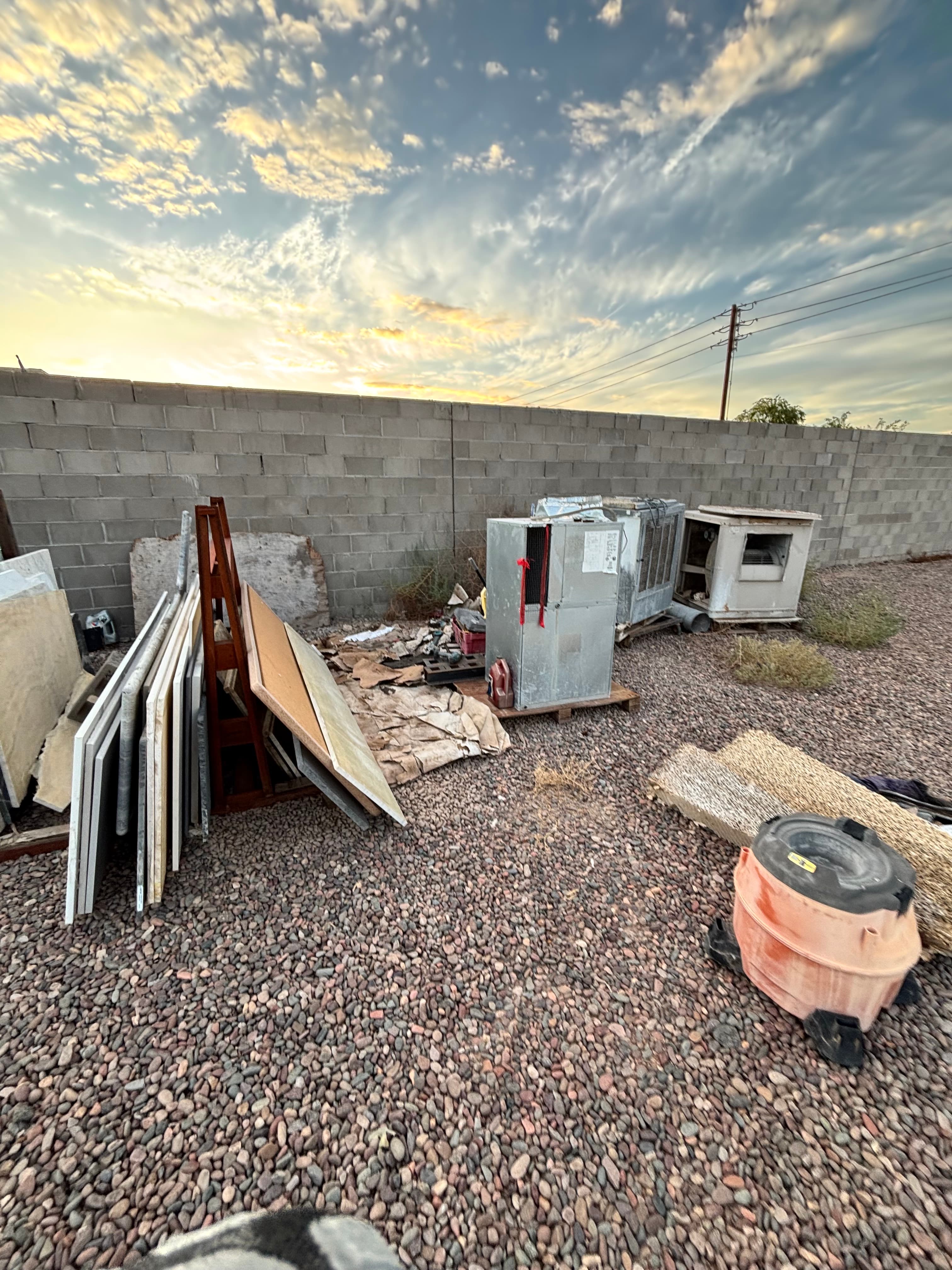 Discarded appliances and building materials in a gravel yard under a dramatic sunset sky.